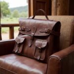 Brown leather messenger bag resting on leather armchair inside wooden cabin with window landscape view