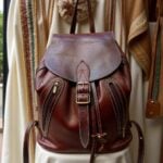 Leather backpack brown on rustic shelf with pottery, featuring hand-stitched details and brass hardware