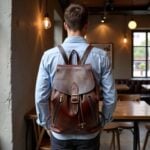 Leather backpack brown displayed on man in cafe, featuring full-grain leather and brass hardware