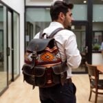 Man wearing dark brown leather kilim backpack with geometric handwoven patterns outdoors near metal gate