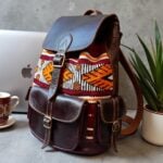 Side view of dark brown leather kilim backpack on workspace desk beside laptop, showing depth and side pocket