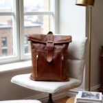Cognac brown leather rolltop backpack resting on a chair by a window, showing buckle closure and front zip pockets