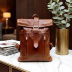 Cognac brown leather rolltop backpack displayed on a marble table with books, featuring hand-stitched seams and buckle closure