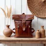 Cognac brown leather rolltop backpack placed on a rustic wooden shelf alongside pottery, showing buckle closure