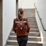 Woman ascending outdoor stairs wearing a cognac brown leather rolltop backpack with buckle closure and front zip pockets