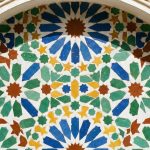 A close-up photograph of an intricate geometric Zellige tile pattern on a wall in a sunlit Moroccan riad courtyard, featuring blue, green, yellow, and red ceramic tiles.