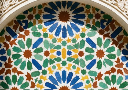 A close-up photograph of an intricate geometric Zellige tile pattern on a wall in a sunlit Moroccan riad courtyard, featuring blue, green, yellow, and red ceramic tiles.