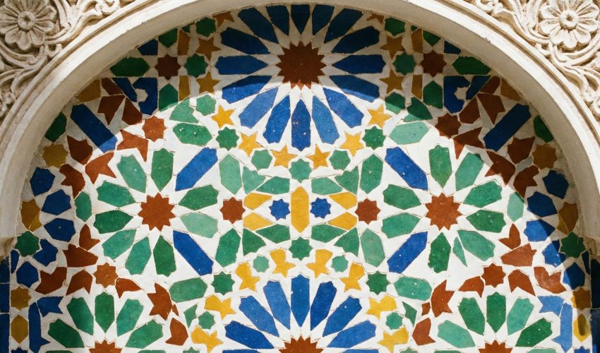 A close-up photograph of an intricate geometric Zellige tile pattern on a wall in a sunlit Moroccan riad courtyard, featuring blue, green, yellow, and red ceramic tiles.