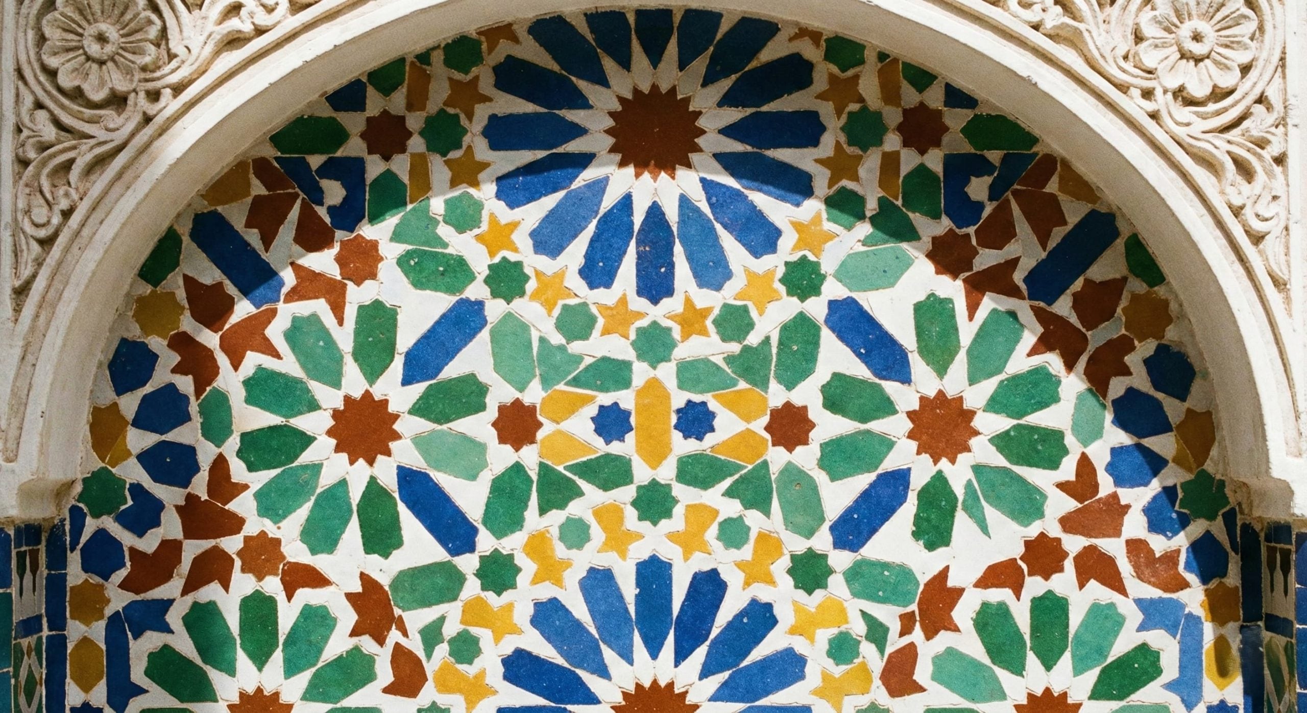 A close-up photograph of an intricate geometric Zellige tile pattern on a wall in a sunlit Moroccan riad courtyard, featuring blue, green, yellow, and red ceramic tiles.