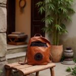Handmade cognac leather backpack with pleated sun design on bamboo bench in rustic courtyard setting