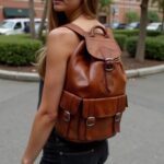 Woman in park with brown leather buckle backpack, close view of brass buckles and hand-stitched details