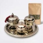 A gleaming hand hammered silver tea container service featuring three footed, lidded bowls on a matching decorative tray, with a red tassel accenting one lid, displayed alongside a kraft paper pouch of loose leaf green tea against a crisp white background.