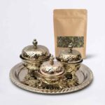 A gleaming hand-embossed silver tea container set featuring three ornate, lidded bowls resting on a matching decorative tray, displayed alongside a kraft paper pouch filled with loose leaf green tea against a clean white background.