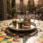 Handmade silver serving tray resting on a colorful mosaic zellige table, featuring a traditional silver teapot and two glasses of steaming mint tea in sunlight.