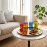 A set of multicolored gold emblem moroccan tea glasses arranged on a traditional round brass tray with sweet Moroccan pastries, placed on a marble coffee table in a bright, modern living room.