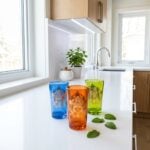 Three vibrant gold emblem moroccan tea glasses in blue, orange, and green resting on a bright white quartz kitchen countertop alongside fresh mint leaves.