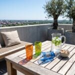 Colorful gold emblem moroccan tea glasses catching the sunlight on a wooden patio table in a sunny outdoor setting, styled with a glass pitcher of mint water and a bowl of sugar cubes.