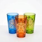 A studio close-up of three bright gold emblem moroccan tea glasses in blue, orange, and green, showcasing their intricate hand-applied golden crest designs against a clean white background.