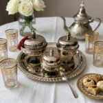 A hand hammered silver tea set styled on a white linen tablecloth with a silver teapot, gold-rimmed glasses, Moroccan cookies, and a vase of white roses.