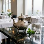 A moroccan teapot hand-engraved in traditional silver is featured on a dark marble table in a luxury restaurant setting, served with fresh mint tea and Moroccan pastries.