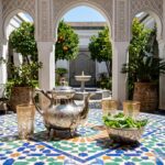 A traditional moroccan teapot hand-engraved is central on a vibrant blue geometric zellige tile table in a peaceful riad courtyard, surrounded by traditional glasses.
