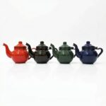 A lineup of four classic Enameled Teapots in varying colors—red, black, green, and blue—displayed in a row against a seamless white studio background.