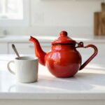 Red moroccan enamel teapot stovetop on white kitchen counter with a ceramic mug and silver spoon