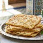 A stack of square, golden-brown Moroccan Msemen flatbreads showing flaky layers, served on a silver tray with a side of honey and a glass of mint tea in a sunlit Moroccan setting.