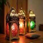 Illuminated engraved brass candle holders casting warm red and green glow with decorative cushions in background