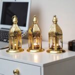 Three engraved brass candle holders with decorative cutouts displayed on white desk beside laptop