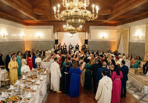 A grand, festive Moroccan wedding hall featuring intricate wall tiles, large brass chandeliers, and elegantly decorated tables in a warm, golden-lit setting.