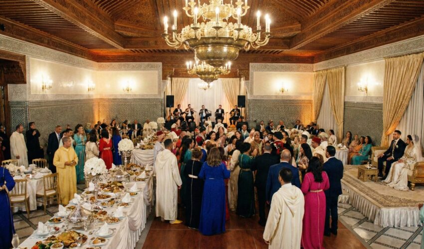 A grand, festive Moroccan wedding hall featuring intricate wall tiles, large brass chandeliers, and elegantly decorated tables in a warm, golden-lit setting.