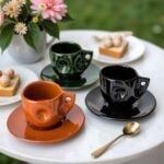 Glossy ceramic coffee cups set in orange, green, and black with decorative swirls and matching saucers on serving tray