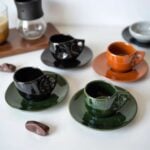 Group of six ceramic coffee cups in black, green, and orange showing artisan swirl design and matching saucers