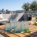 Sunlit light blue beldi glasses adorned with white diamond designs resting on a wooden patio table, styled with a lemon water pitcher and fresh mint on a sunny rooftop terrace.