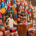 A colorful display of Moroccan leather goods in a souk stall, including hanging babouche slippers in various colors, embossed bags, and poufs.