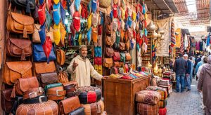 A colorful display of Moroccan leather goods in a souk stall, including hanging babouche slippers in various colors, embossed bags, and poufs.