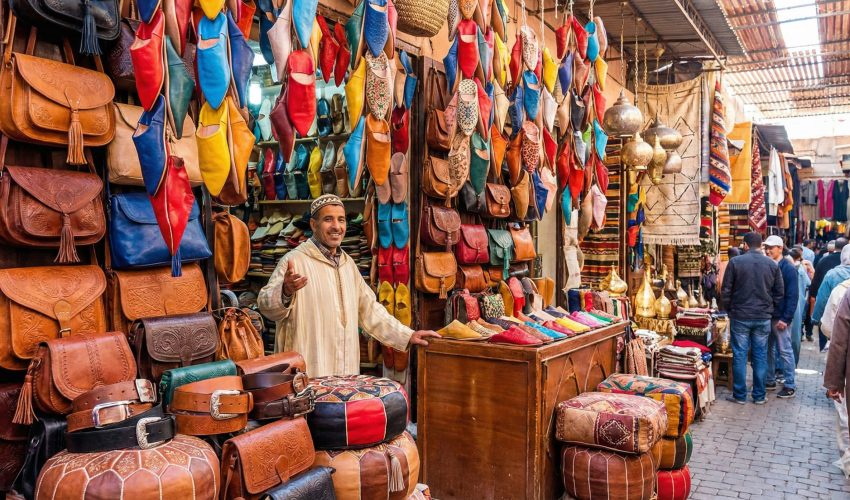 A colorful display of Moroccan leather goods in a souk stall, including hanging babouche slippers in various colors, embossed bags, and poufs.