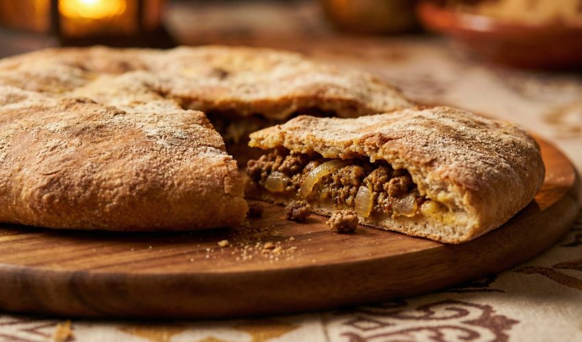 Sliced Moroccan Berber pizza on a wooden board showing the seasoned ground lamb and onion filling inside the golden crust