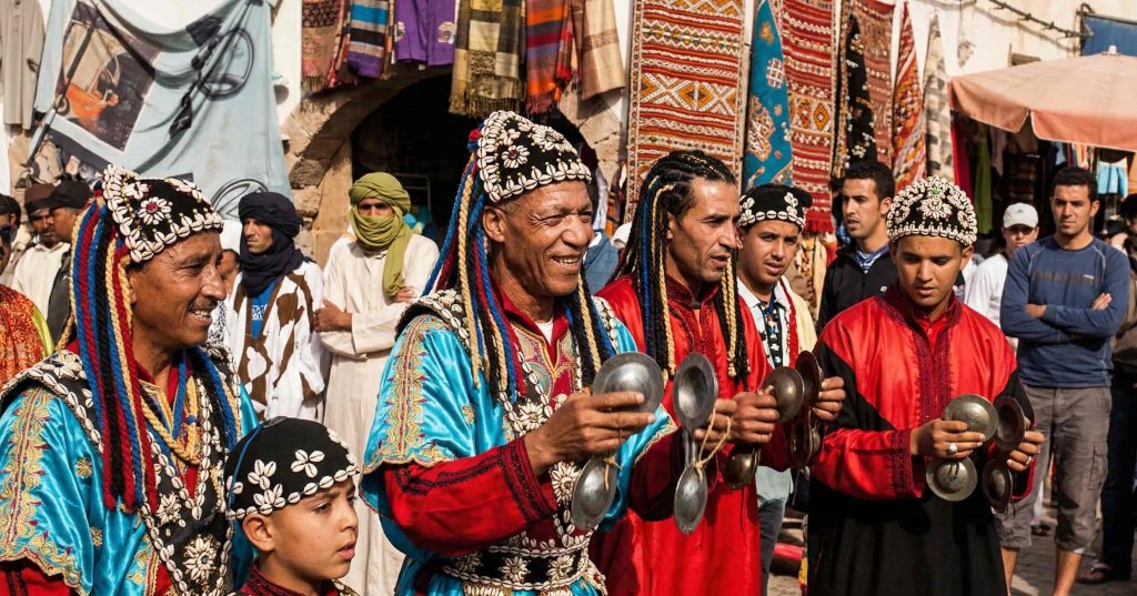The Enchanting Tapestry: Examining What Makes Morocco Famous A group of traditional Gnawa musicians performing in a lively outdoor Moroccan market. They are wearing vibrant blue and red robes heavily adorned with white cowrie shells, along with matching ornate caps featuring long, colorful tassels. The smiling men are playing large iron castanets, known as qraqeb, while a young boy in similar attire stands beside them. In the background, a crowd of onlookers gathers near shops displaying colorful hanging textiles and carpets.