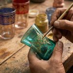A close-up photograph of an artisan’s hands carefully painting gold filigree patterns on a teal-colored Moroccan tea glass in a workshop.