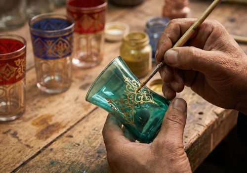 A close-up photograph of an artisan’s hands carefully painting gold filigree patterns on a teal-colored Moroccan tea glass in a workshop.