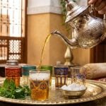 Moroccan mint tea being poured from a silver teapot into colorful hand-painted glasses on an ornate brass tray with fresh mint