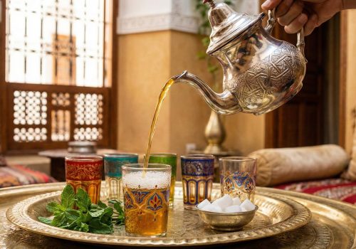 Moroccan mint tea being poured from a silver teapot into colorful hand-painted glasses on an ornate brass tray with fresh mint