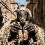 A close-up photograph of a Boujloud festival participant wearing traditional goat skins, a horned mask, and charcoal face paint in a sunlit Moroccan alley.