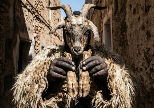 A close-up photograph of a Boujloud festival participant wearing traditional goat skins, a horned mask, and charcoal face paint in a sunlit Moroccan alley.