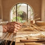 A close-up photograph of an artisan’s hands weaving natural fibers in a bright, modern studio, surrounded by sustainable handmade Moroccan pottery and textiles, symbolizing the preservation of tradition.