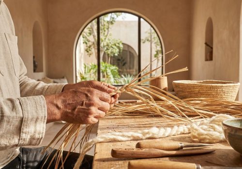A close-up photograph of an artisan’s hands weaving natural fibers in a bright, modern studio, surrounded by sustainable handmade Moroccan pottery and textiles, symbolizing the preservation of tradition.