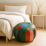Colorful leather pouf ottoman placed at the foot of a minimalist bed in a bright, modern bedroom.