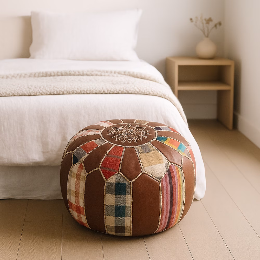 Colorful patchwork round leather pouf ottoman placed in a soft minimalist bedroom beside a natural wood nightstand.